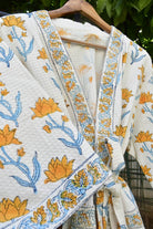 A white cotton kimono robe with a yellow and blue floral block print pattern, hanging on a wooden hanger against a background of green foliage.