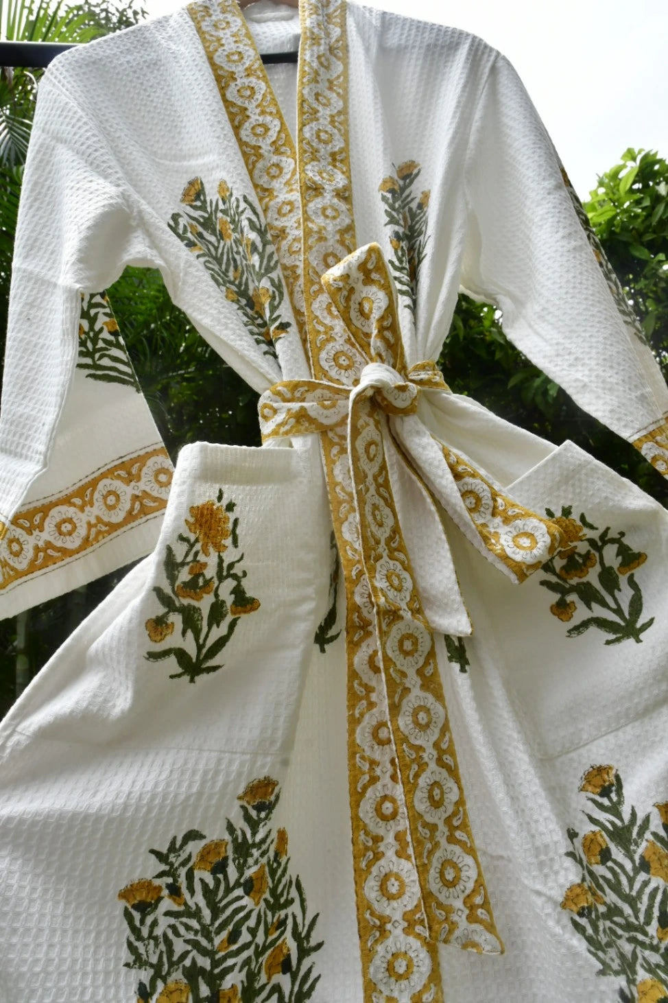 A white waffle-knit robe with yellow and green floral embroidery and patterned borders, tied with a matching belt, shown outdoors against a background of green foliage.