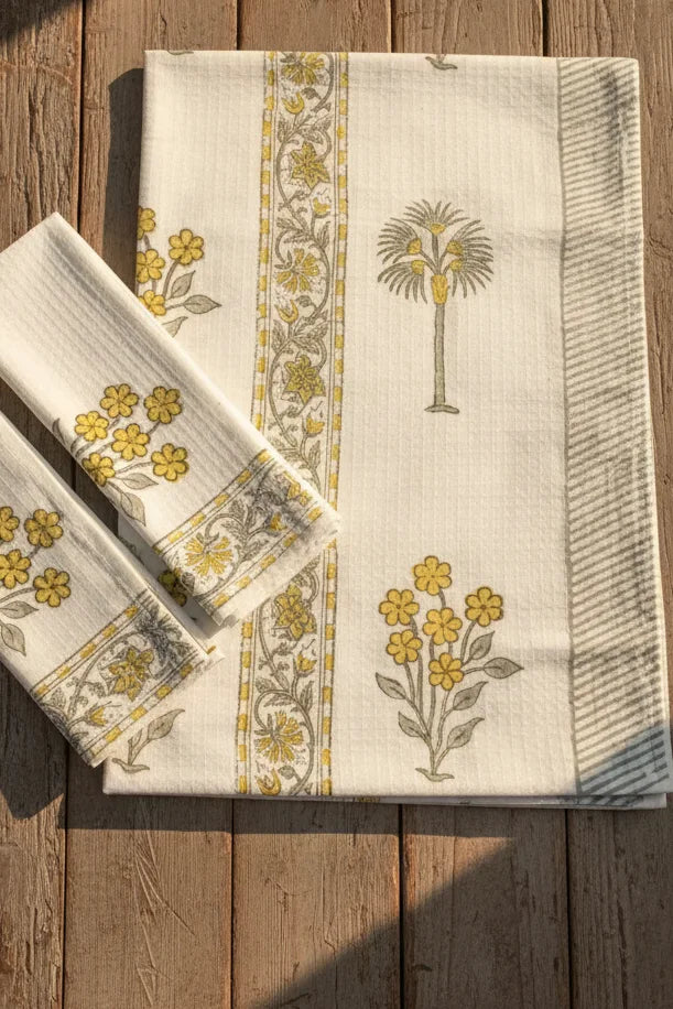 A folded cream-colored cotton tablecloth with yellow floral patterns and a central vertical border, displayed on a rustic wooden plank surface.