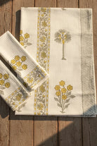 A folded cream-colored cotton tablecloth with yellow floral patterns and a central vertical border, displayed on a rustic wooden plank surface.