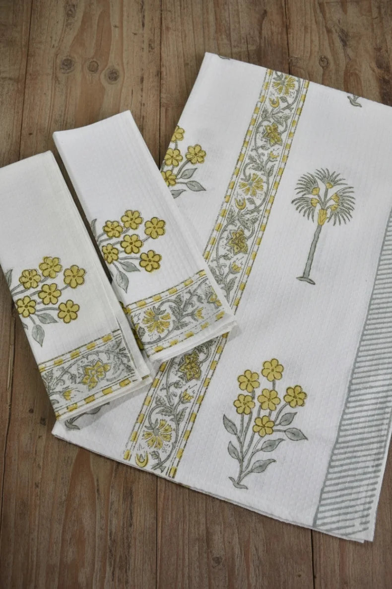 A set of white cotton kitchen towels with yellow floral prints and gray-green botanical borders, arranged on a rustic wooden surface.