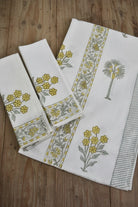 A set of white cotton kitchen towels with yellow floral prints and gray-green botanical borders, arranged on a rustic wooden surface.