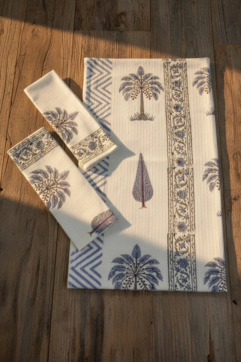 A set of cream-colored cotton towel set  including a large placemat and two matching napkins, featuring a blue and brown palm tree and geometric border print, arranged on a rustic wooden table with warm sunlight.