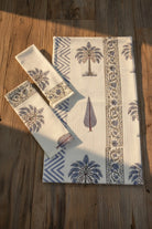 A set of cream-colored cotton towel set  including a large placemat and two matching napkins, featuring a blue and brown palm tree and geometric border print, arranged on a rustic wooden table with warm sunlight.