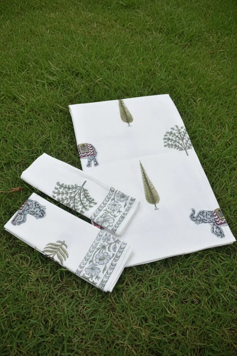 A set of white cotton towels with hand-painted elephant and tree motifs, arranged on a green grass lawn.