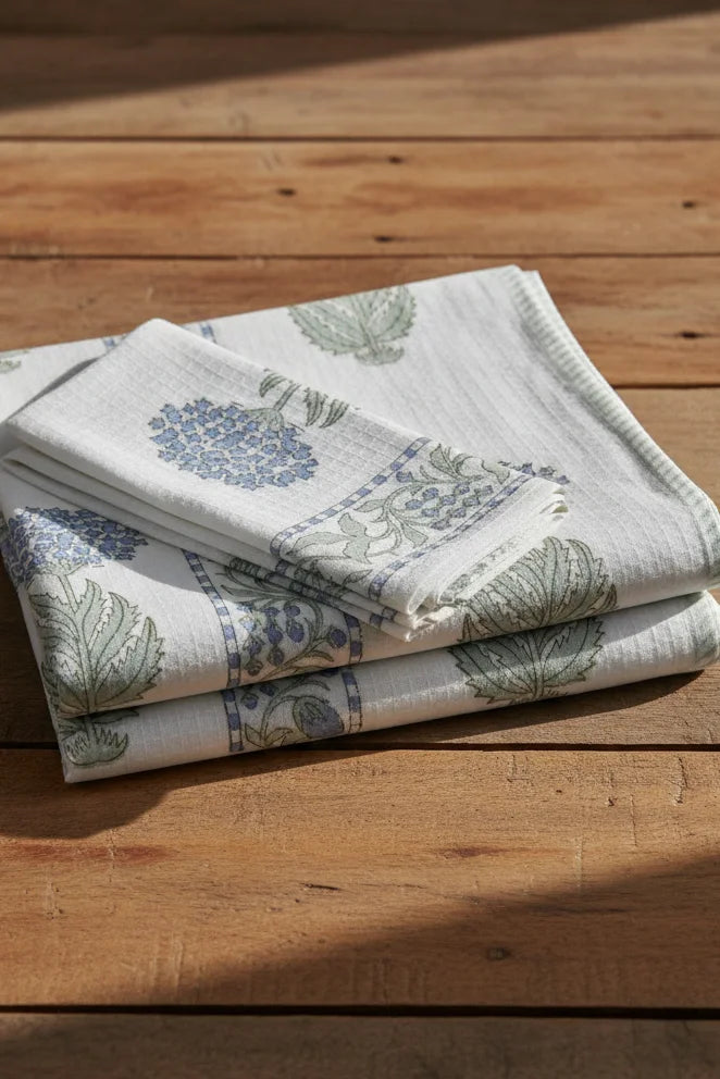 A stack of folded cotton towels with a blue and green botanical block print pattern, displayed on a rustic wooden surface.