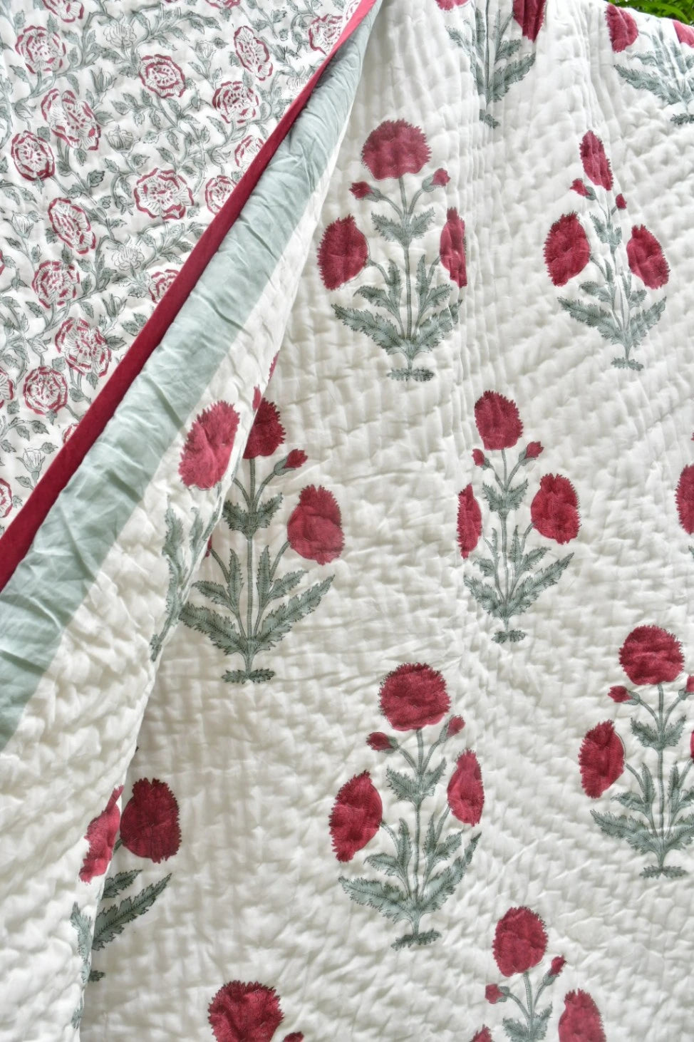 A close-up of a quilted textile with a white base, red floral motifs, and pale green stems, plus a folded section showing a red border and a second patterned side.