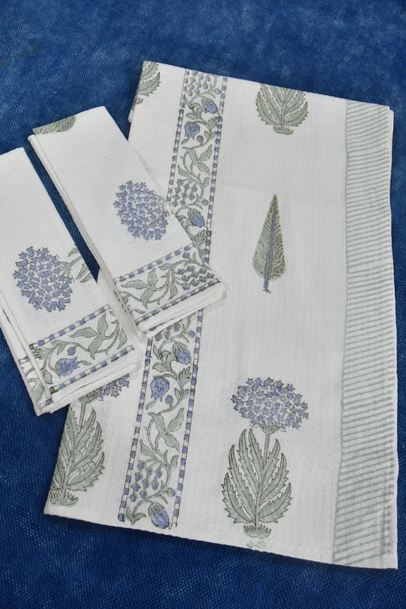 A folded white cotton napkin set with blue and green floral block prints, displayed on a textured blue background.
